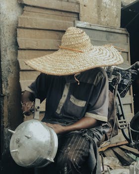 Man in straw hat crafting a metal pot in outdoor market, Kaduna, Nigeria.
